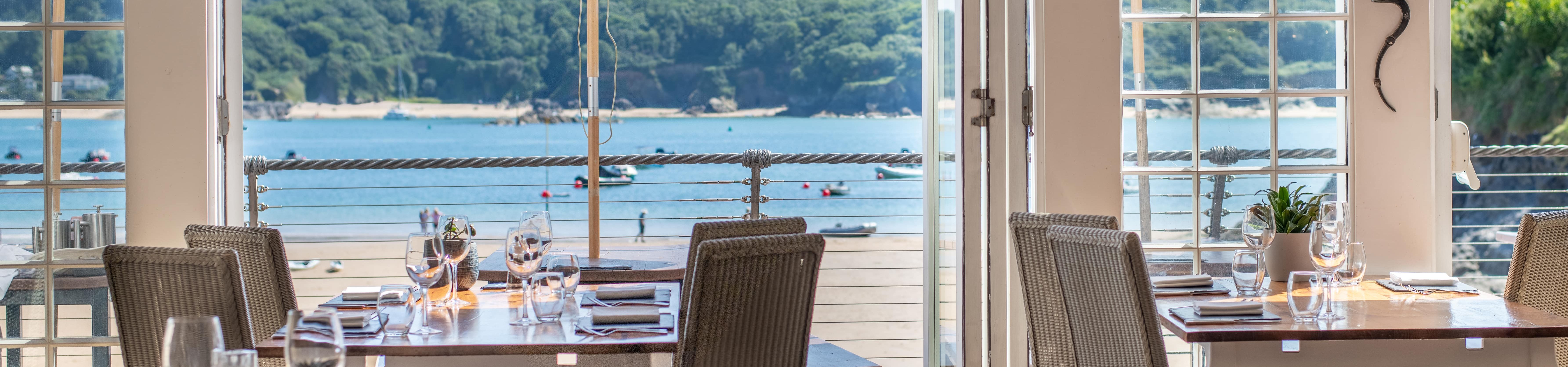 Seaside restaurant interior with tables set for dining, open doors, and a scenic view of the beach and ocean outside.