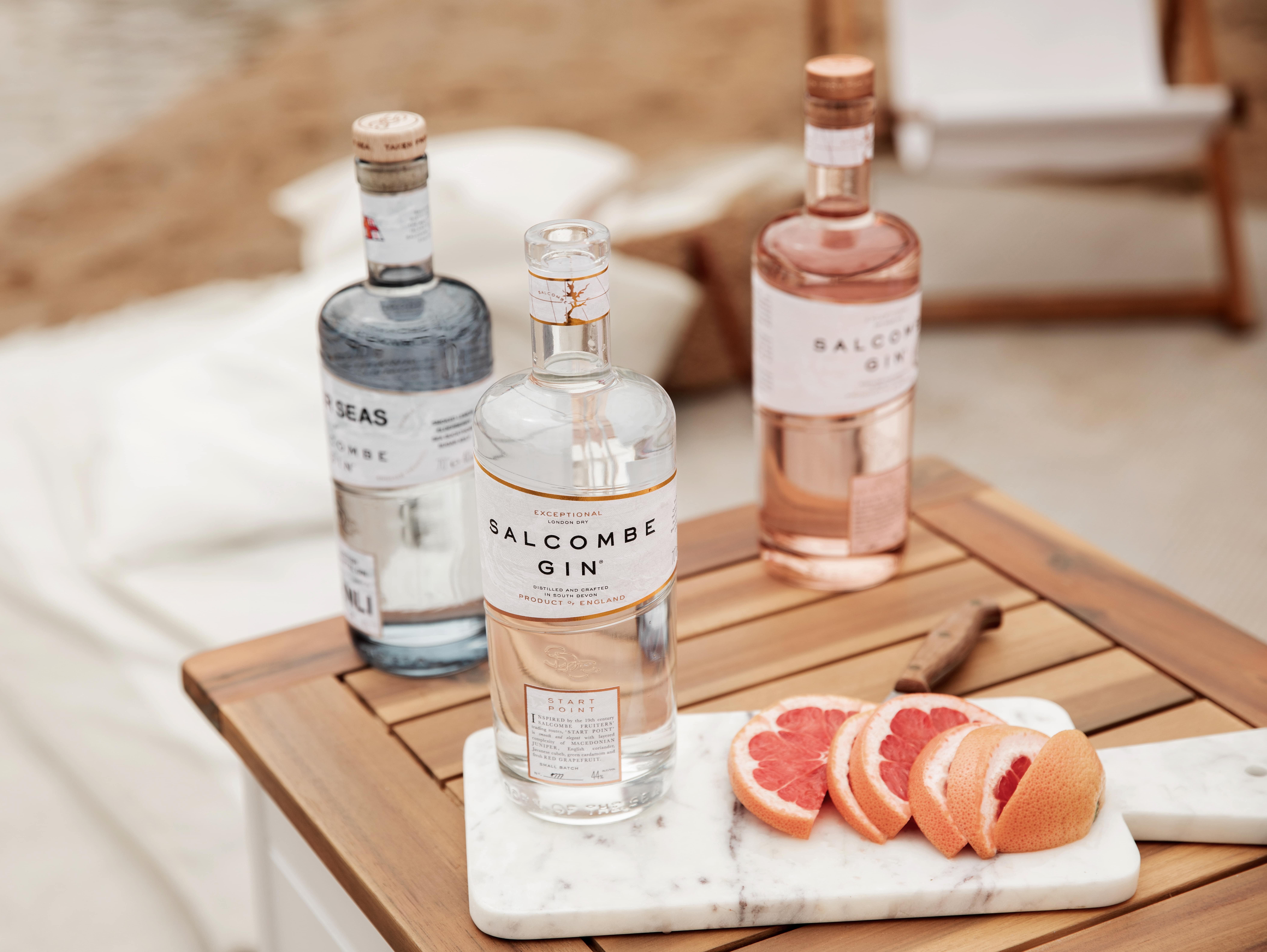 Three bottles of Salcombe Gin on a wooden table, next to sliced grapefruit on a marble board.