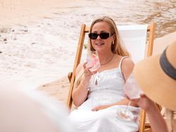 Woman relaxing on a beach chair, wearing sunglasses and a white dress, holding a cocktail, with another person beside her.
