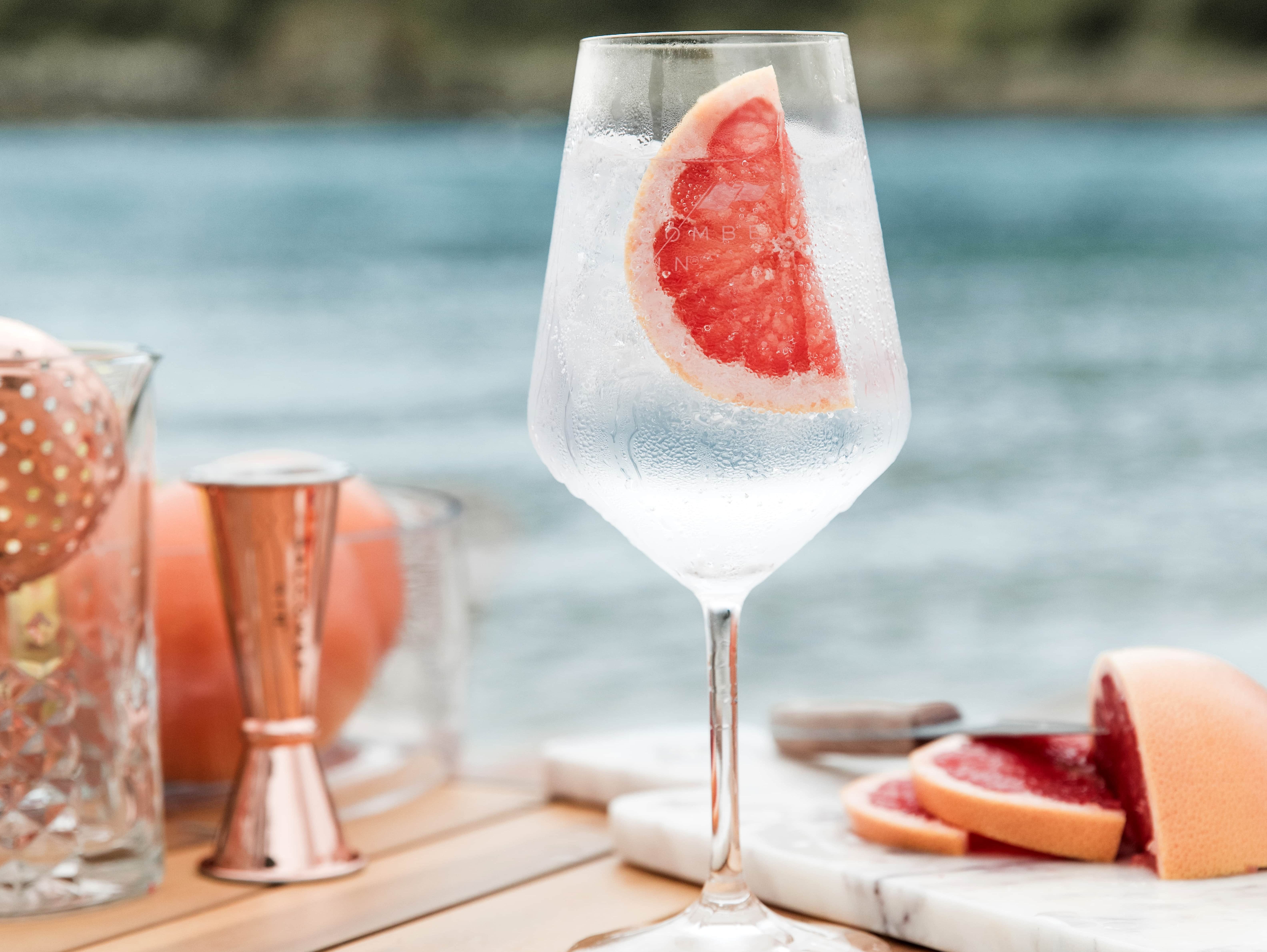 A glass of cocktail garnished with a grapefruit slice, sitting on a wooden table by the water.