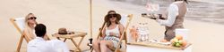People relaxing on a beach under a Salcombe Gin umbrella, being served drinks by a staff member.