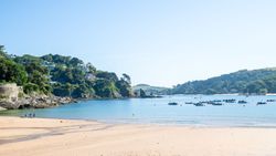 Sandy beach with clear blue water, small boats anchored offshore, and green hills with houses in the background under a bright blue sky.