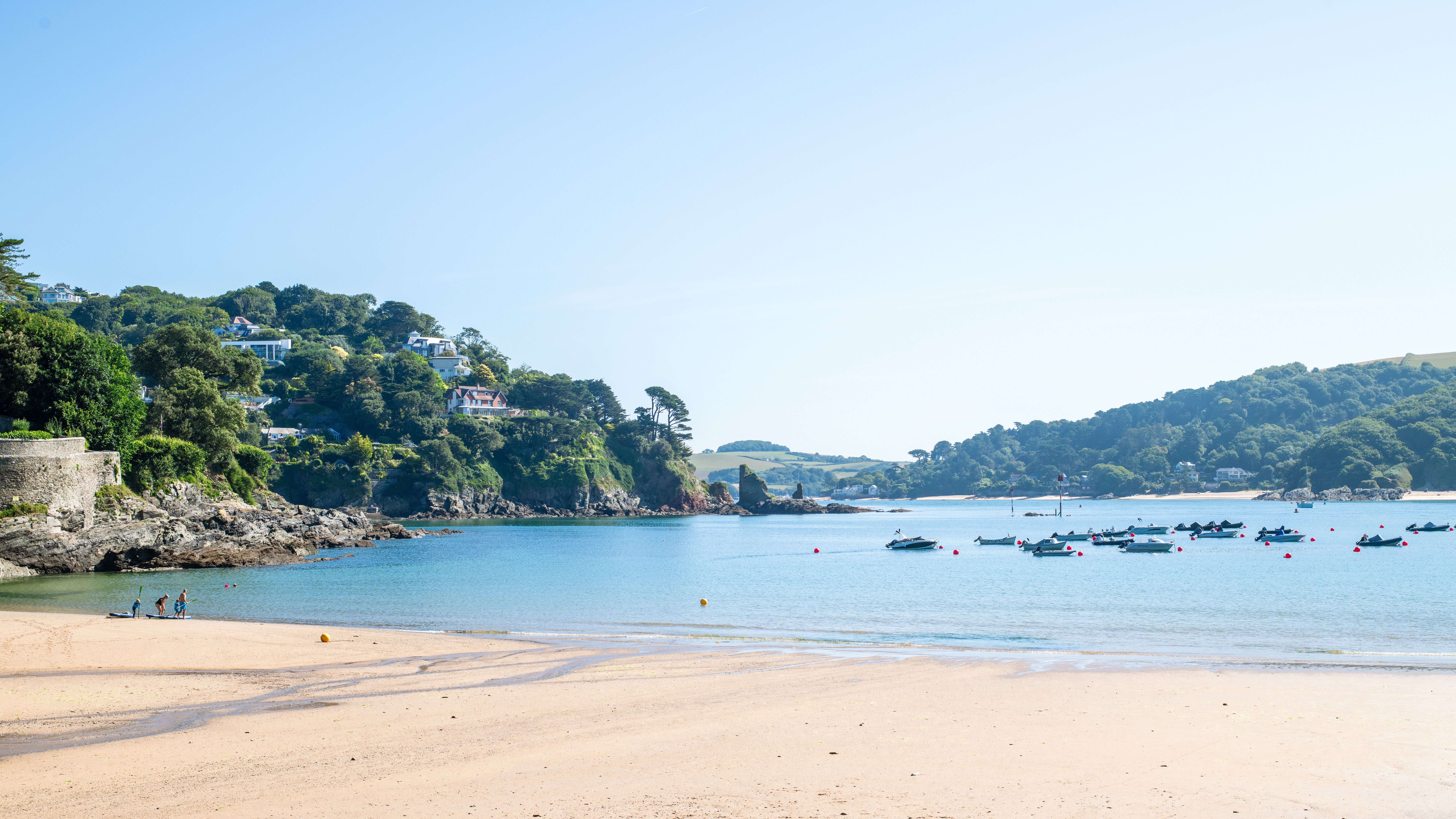 Sandy beach with clear blue water, small boats anchored offshore, and green hills with houses in the background under a bright blue sky.