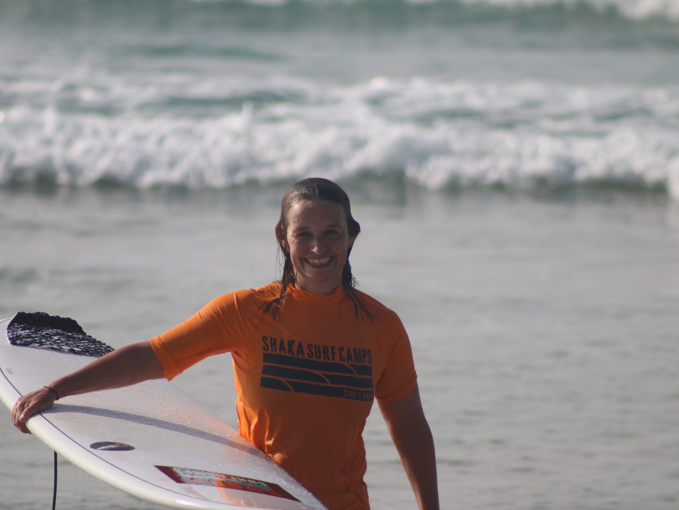 Smiling surfer in an orange rash vest holding a surfboard in shallow sea water with waves behind