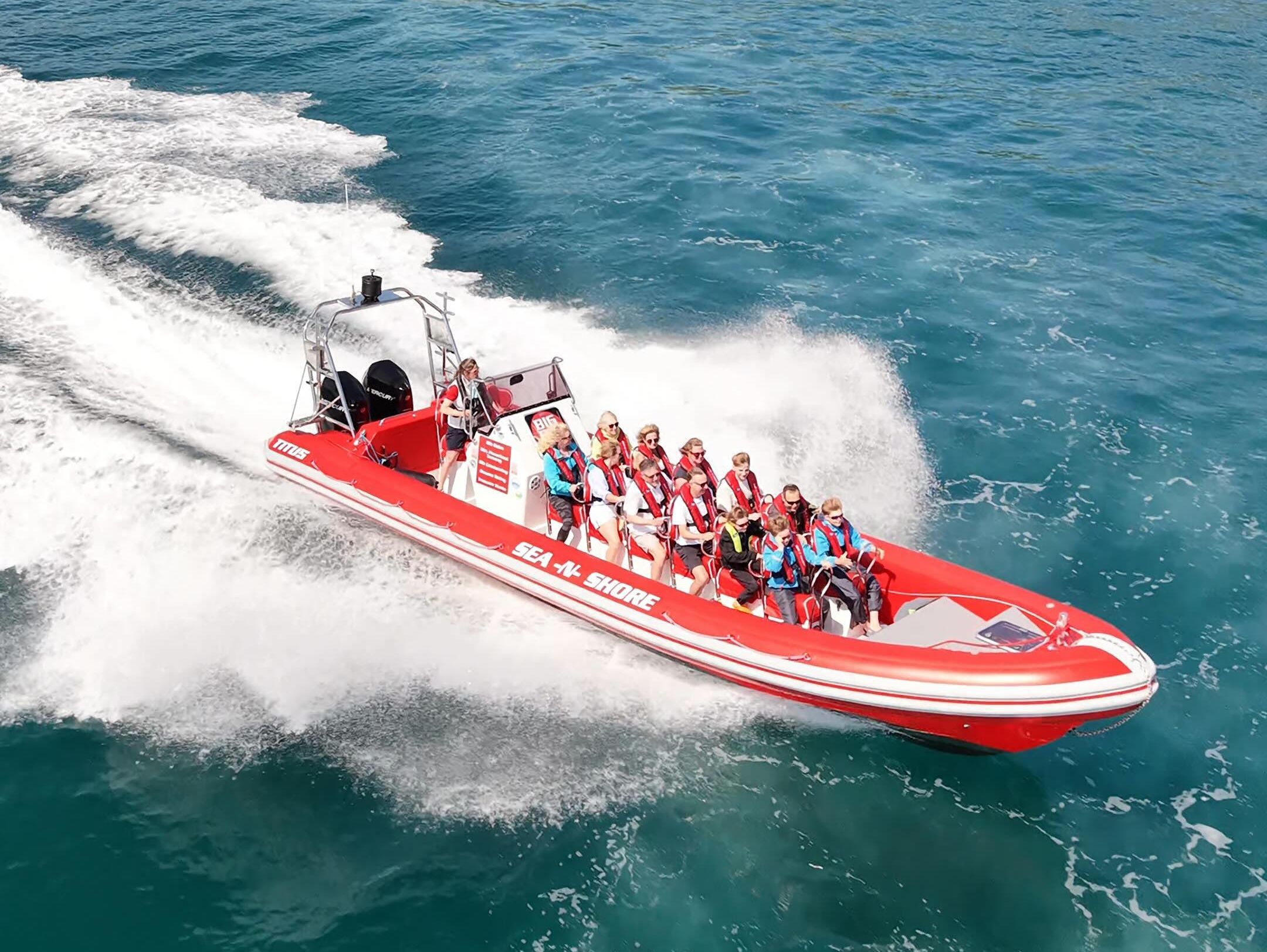 Red RIB speedboat carrying passengers in lifejackets speeding across turquoise sea near a rocky, green coastline