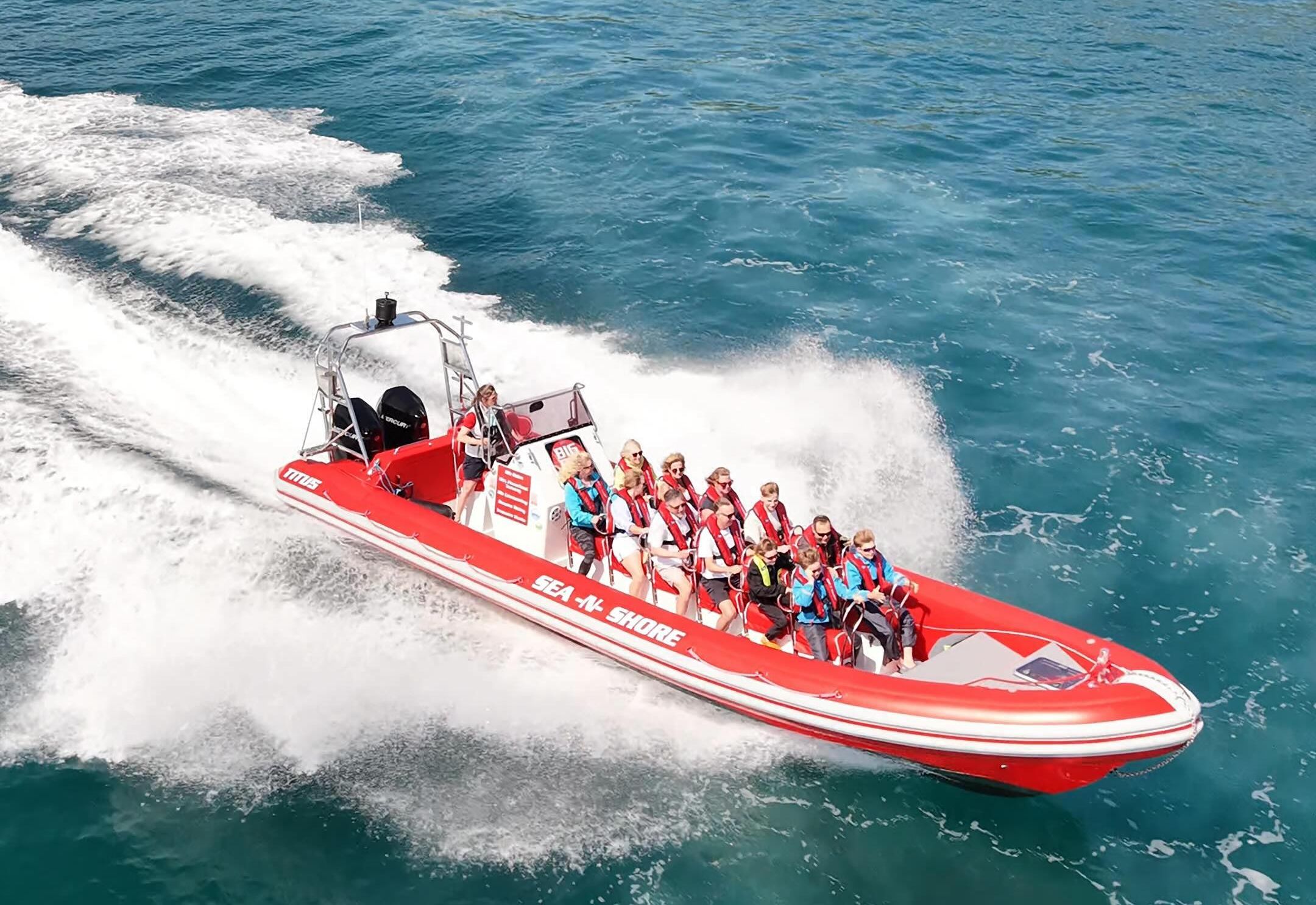 Red RIB speedboat carrying passengers in lifejackets speeding across turquoise sea near a rocky, green coastline
