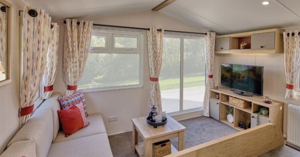 Cozy living room with a beige sectional sofa, patterned curtains, a wooden coffee table, a modern TV on a wooden entertainment unit, and large windows with a view of greenery outside.