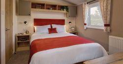 Modern cozy bedroom with a double bed featuring red and white bedding, bedside tables, wall-mounted reading lights, shelves with decorative items, and a window with patterned curtains.