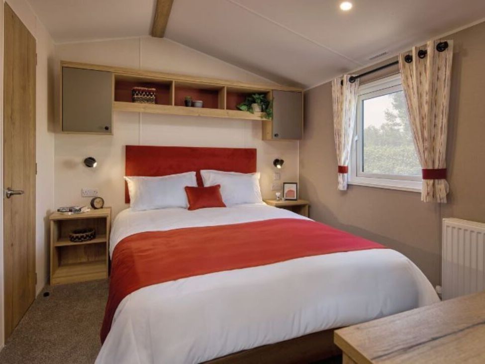 Modern cozy bedroom with a double bed featuring red and white bedding, bedside tables, wall-mounted reading lights, shelves with decorative items, and a window with patterned curtains.