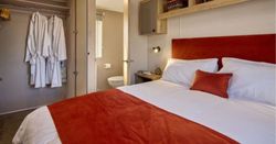 Cozy bedroom with orange and white bedding, open wardrobe with white bathrobes, bedside table, overhead shelves, and a view into an adjacent bathroom.