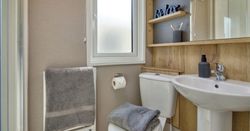 Small modern bathroom with a white toilet, pedestal sink, wooden shelves with toiletries, a towel radiator holding gray towels, and a frosted window.