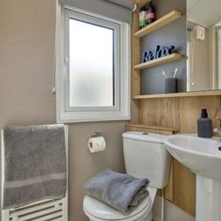 Small modern bathroom with a white toilet, pedestal sink, wooden shelves with toiletries, a towel radiator holding gray towels, and a frosted window.