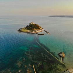 Aerial view of a small island with a historic castle, connected to the mainland by a causeway over clear blue-green waters.