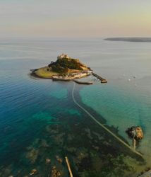 Aerial view of a small island with a historic castle, connected to the mainland by a causeway over clear blue-green waters.