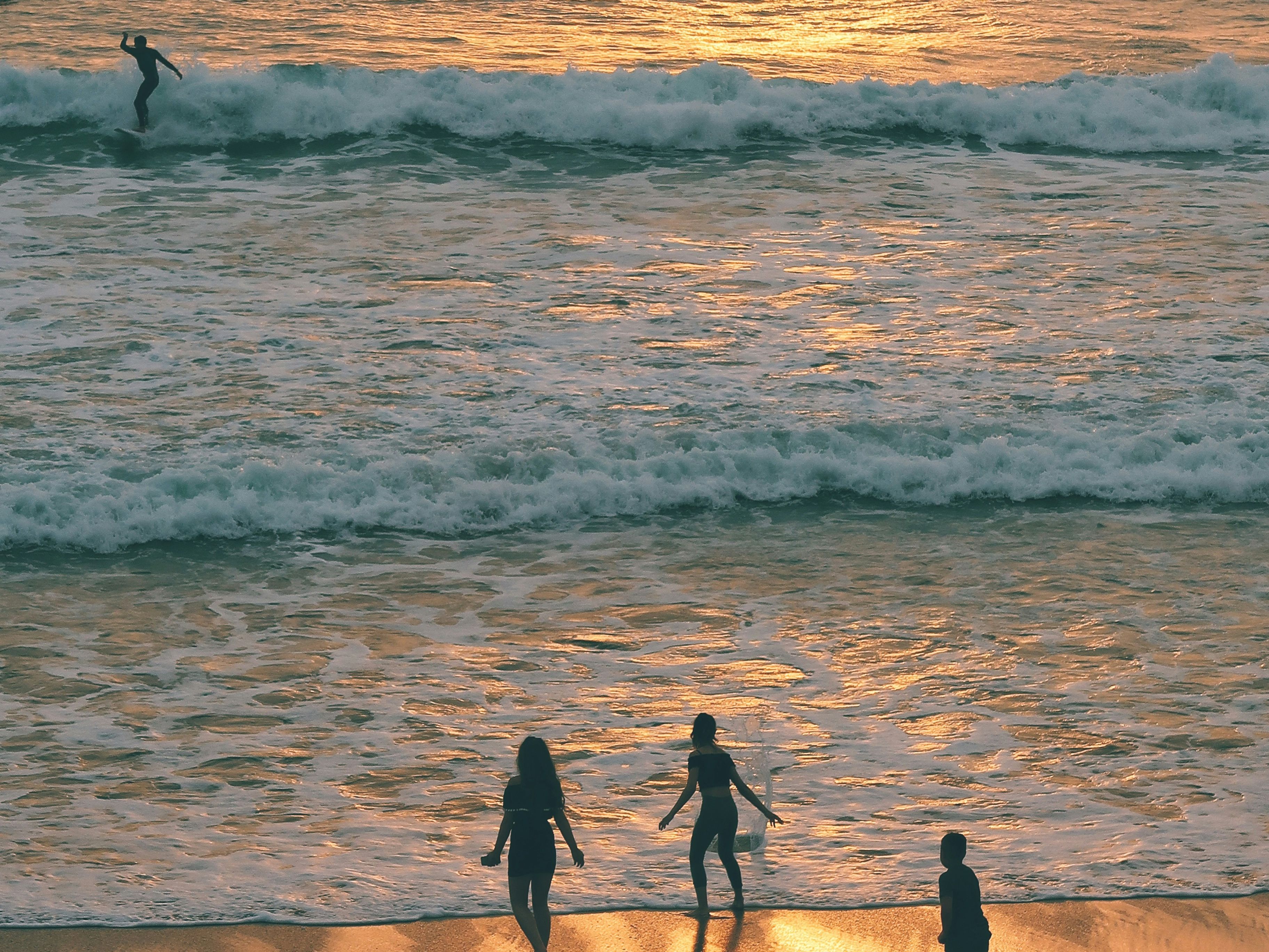 Silhouettes of three people walking on the beach and a surfer riding a wave in the ocean at sunset.
