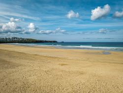 Wide sandy beach with gentle waves, clear blue sky, and a few clouds. Buildings and green hills are visible in the distance along the coastline.