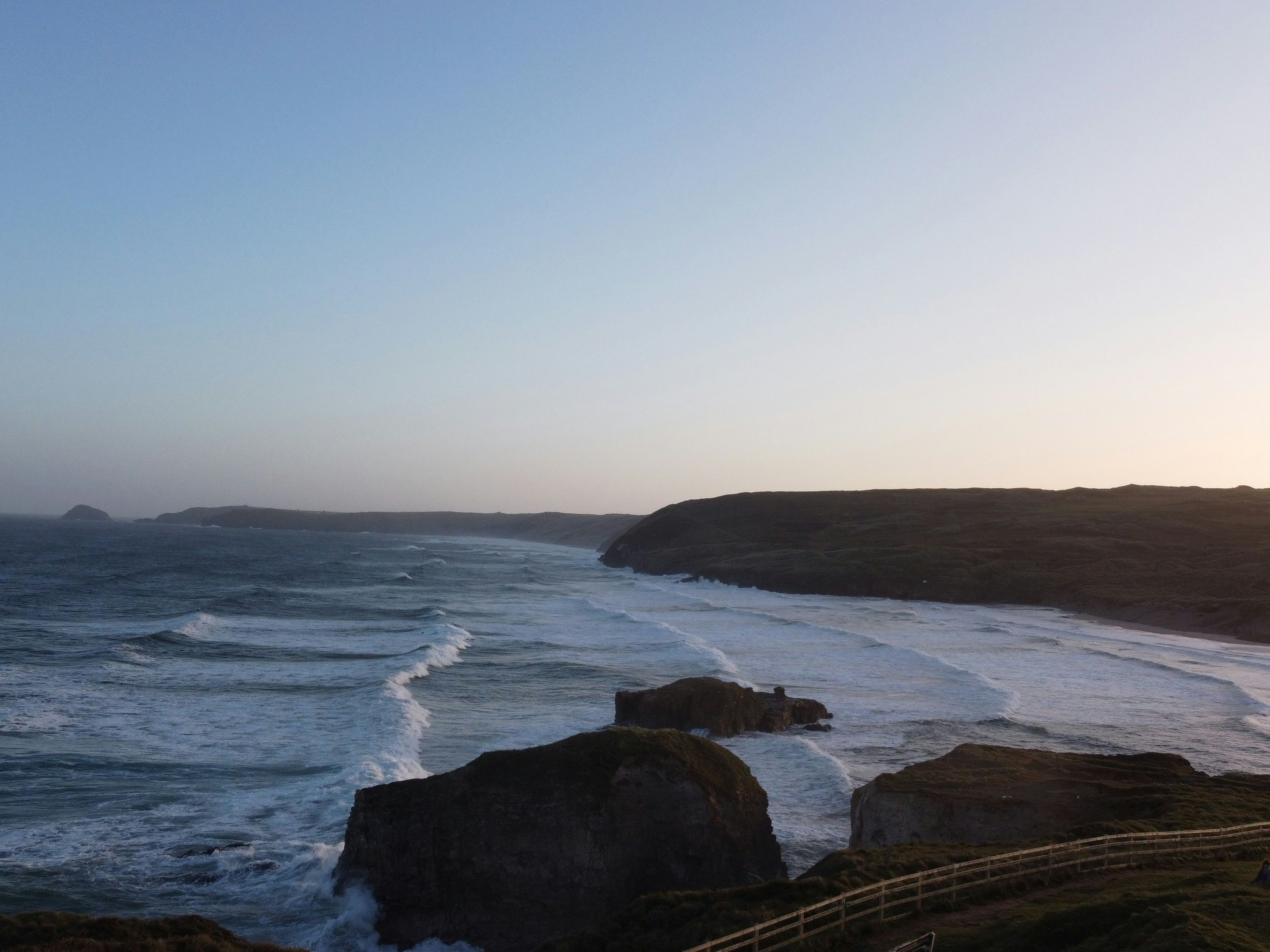 Coastal landscape at sunset with waves crashing on rocks and cliffs, a grassy area with fences in the foreground, and hills in the distance.