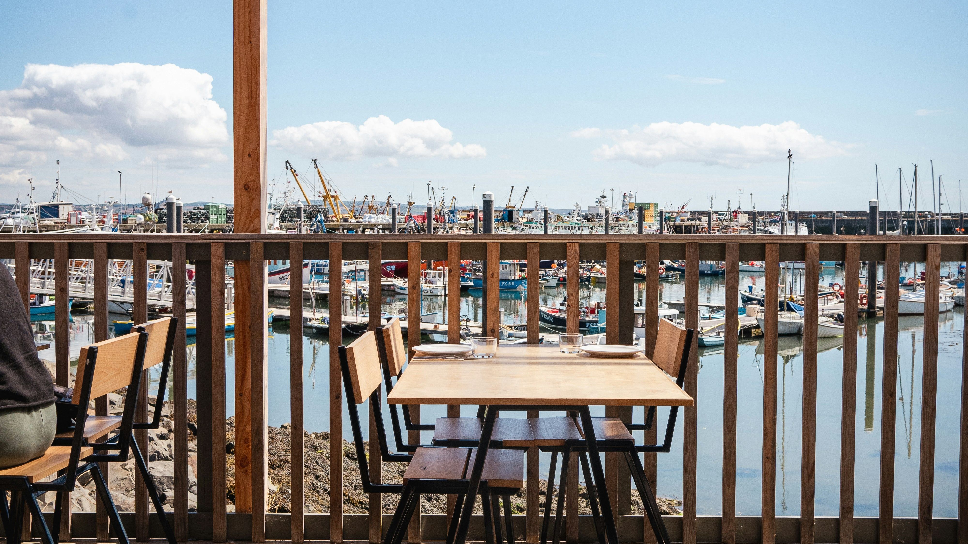 Outdoor seating area with wooden tables and chairs under a pergola, overlooking a marina filled with boats on a sunny day.