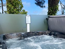 A bubbling outdoor hot tub with swirling water, surrounded by privacy glass panels and trees, under a blue sky. A sign with rules or instructions is posted on the fence behind the hot tub.