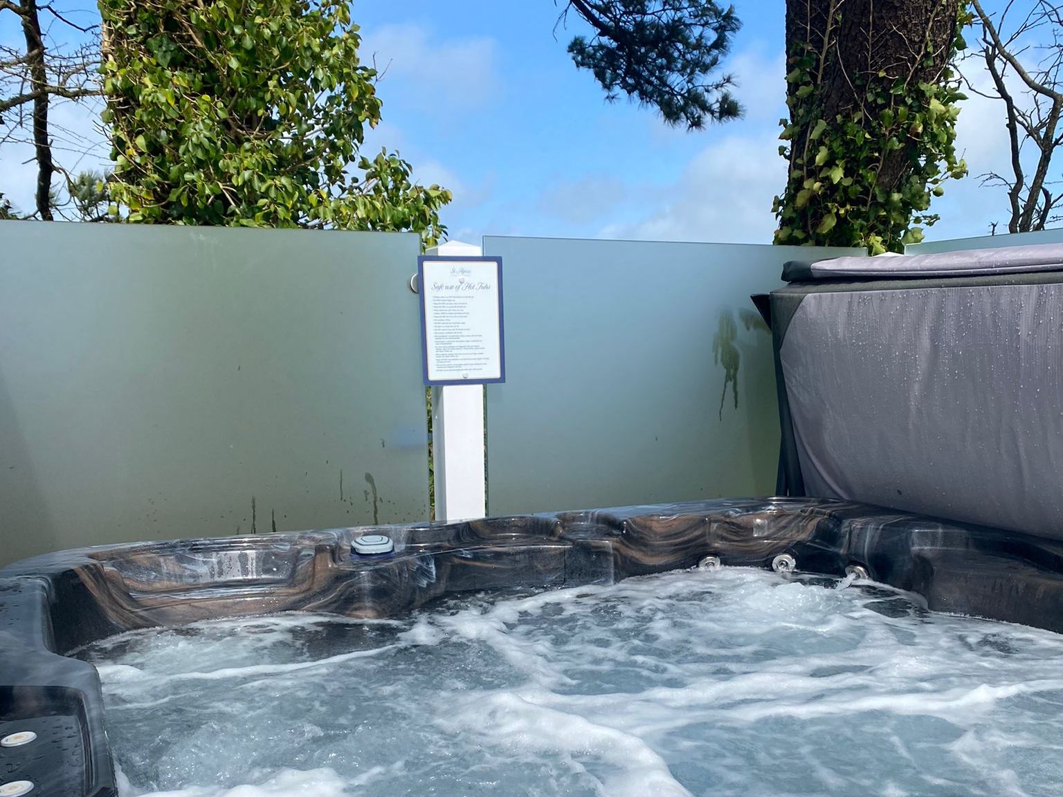 A bubbling outdoor hot tub with swirling water, surrounded by privacy glass panels and trees, under a blue sky. A sign with rules or instructions is posted on the fence behind the hot tub.