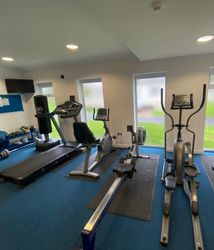 Small gym room with various fitness equipment including treadmills, an elliptical machine, stationary bikes, dumbbells, kettlebells, a TV, and blue carpet flooring.