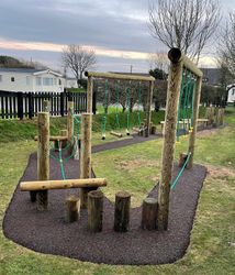 Outdoor wooden playground equipment with balance beams, stepping logs, and ropes, set on a grassy area with mobile homes and trees in the background.