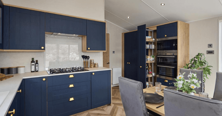 Modern kitchen with navy blue cabinets, gold handles, a herringbone wood floor, integrated appliances, and a dining table set with flowers and grey upholstered chairs.