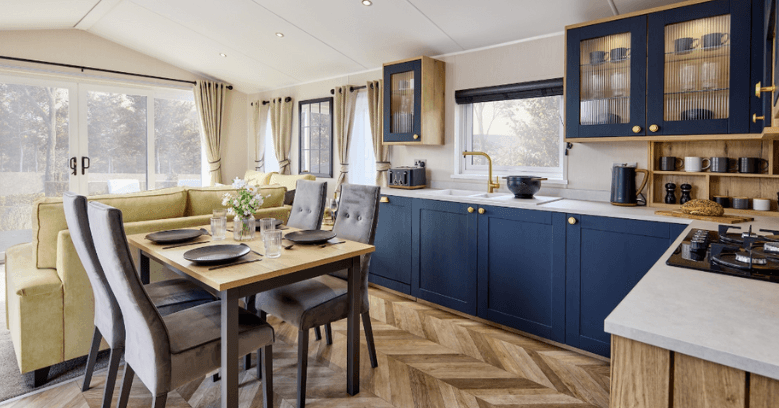 Modern open-plan kitchen and dining area with blue cabinets, wooden accents, herringbone flooring, a yellow sofa, and large windows with beige curtains letting in natural light.