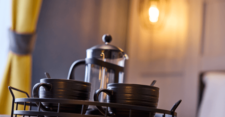 Close-up of a black tray with two black coffee cups and a French press on a bed, with yellow curtains and a blurred wall lamp in the background.