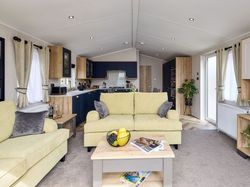 Modern open-plan living room and kitchen with light yellow sofas, coffee table with fruit and magazines, wooden cabinetry, and large windows with curtains.