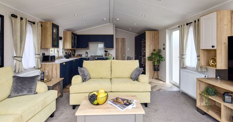 Modern open-plan living room and kitchen with light yellow sofas, coffee table with fruit and magazines, wooden cabinetry, and large windows with curtains.