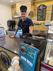A smiling barista wearing a black chef hat and apron standing behind the counter of a coffee shop with a menu and coffee machine visible.