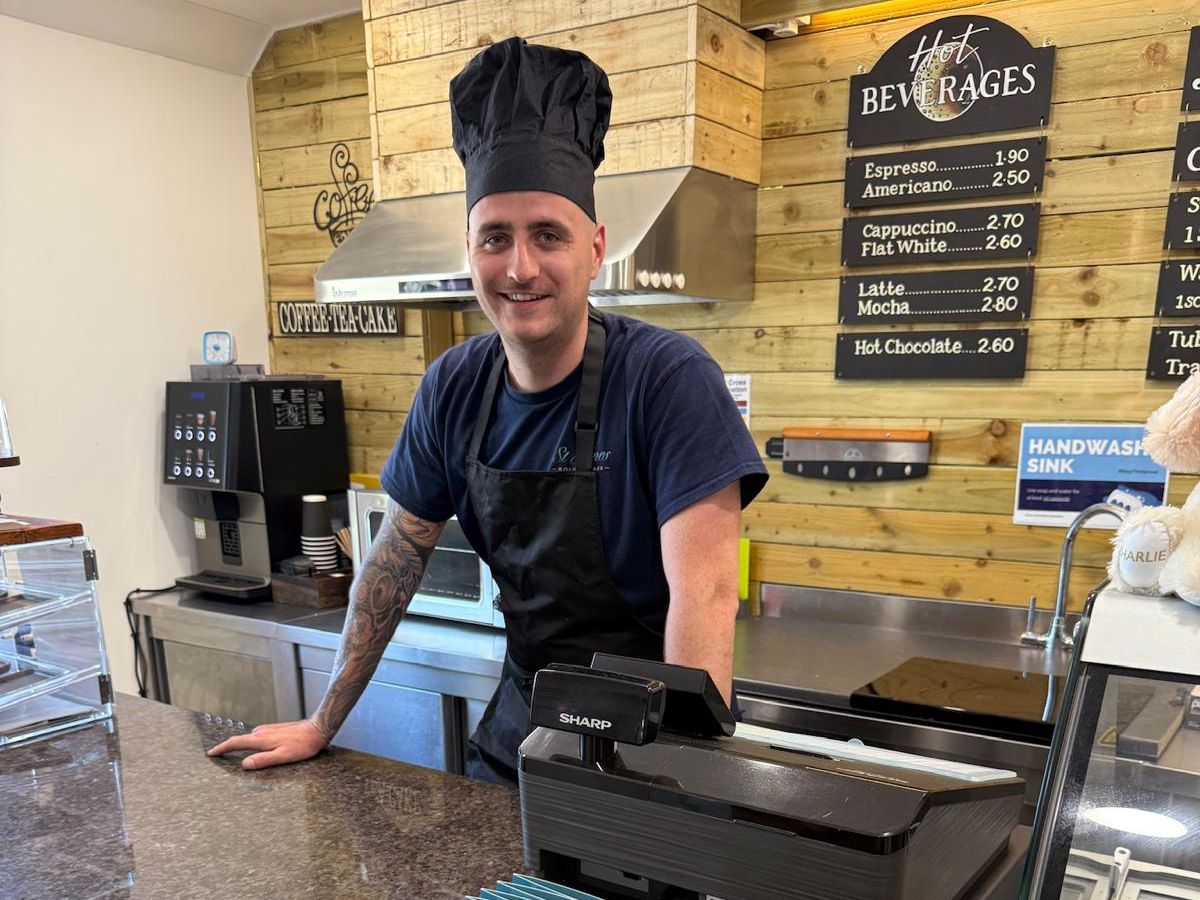 A smiling barista wearing a black chef hat and apron standing behind the counter of a coffee shop with a menu and coffee machine visible.