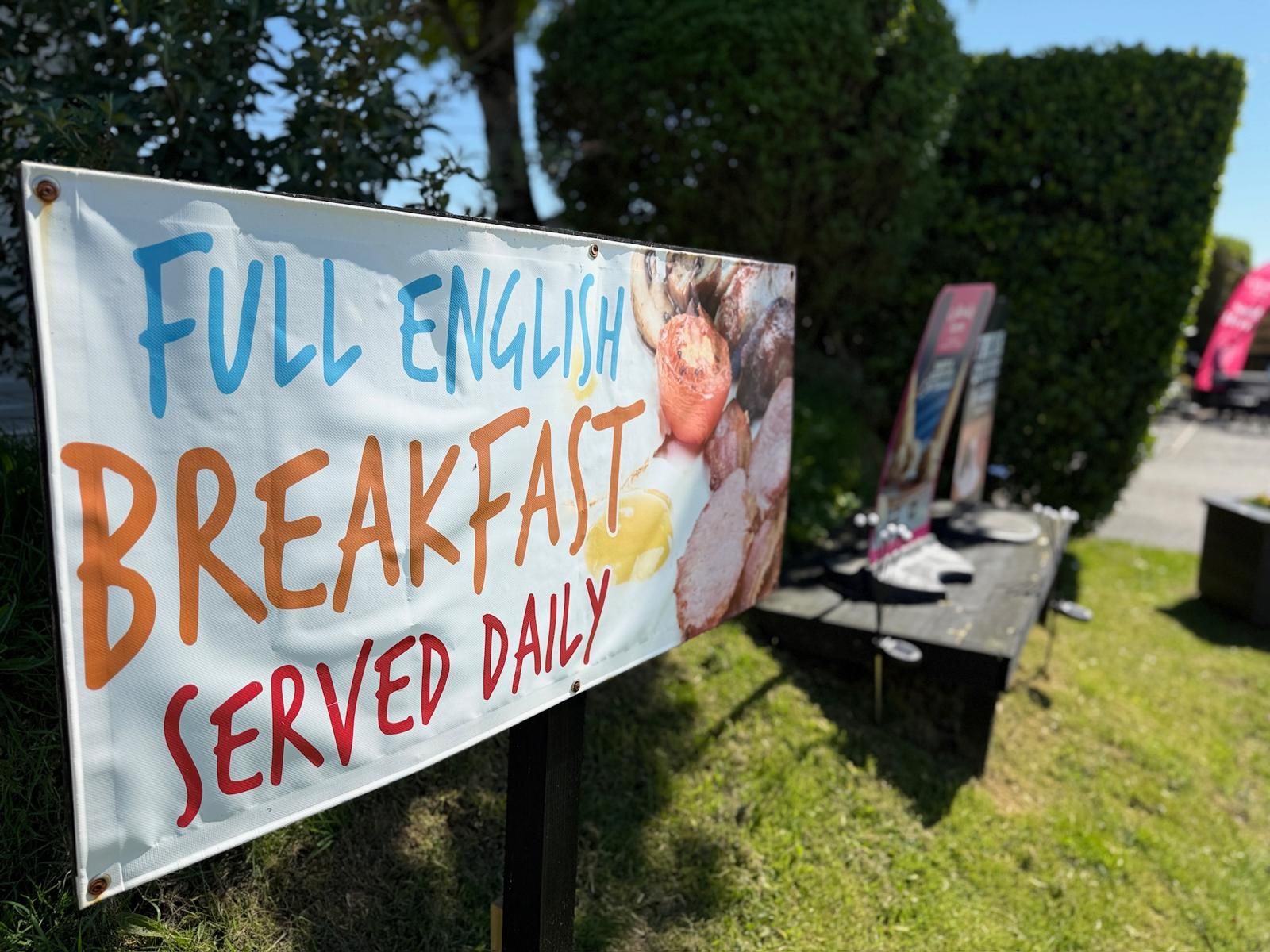 A banner outdoors advertising 'Full English Breakfast Served Daily' with images of breakfast food including eggs, sausages, and bacon.