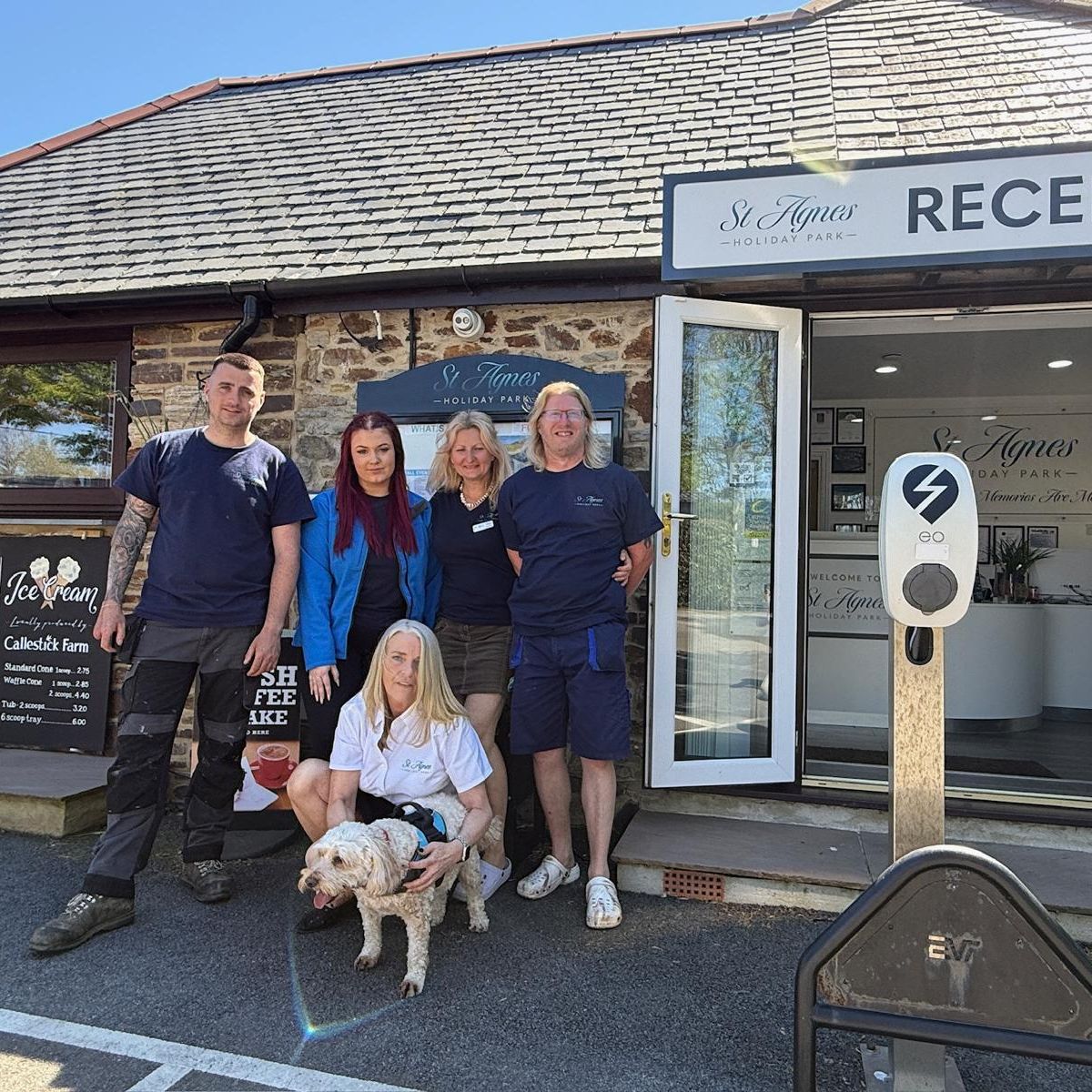 Group of five people and a dog standing outside the reception of St Agnes Holiday Park, with a signboard displaying pizza and ice cream menu on the left.