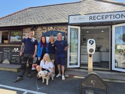 Group of five people and a dog standing outside the reception of St Agnes Holiday Park, with a signboard displaying pizza and ice cream menu on the left.