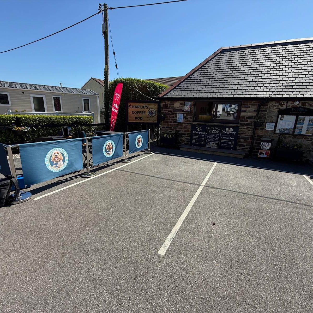 Outdoor seating area in front of a small coffee shop called Charlie's Coffee Pod, with a signboard and menu on display, located next to a stone building on a sunny day.