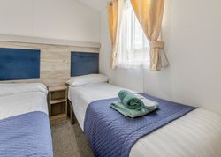 A tidy twin bedroom with two single beds, blue bed runners, folded towels on one bed, and a window with yellow curtains.