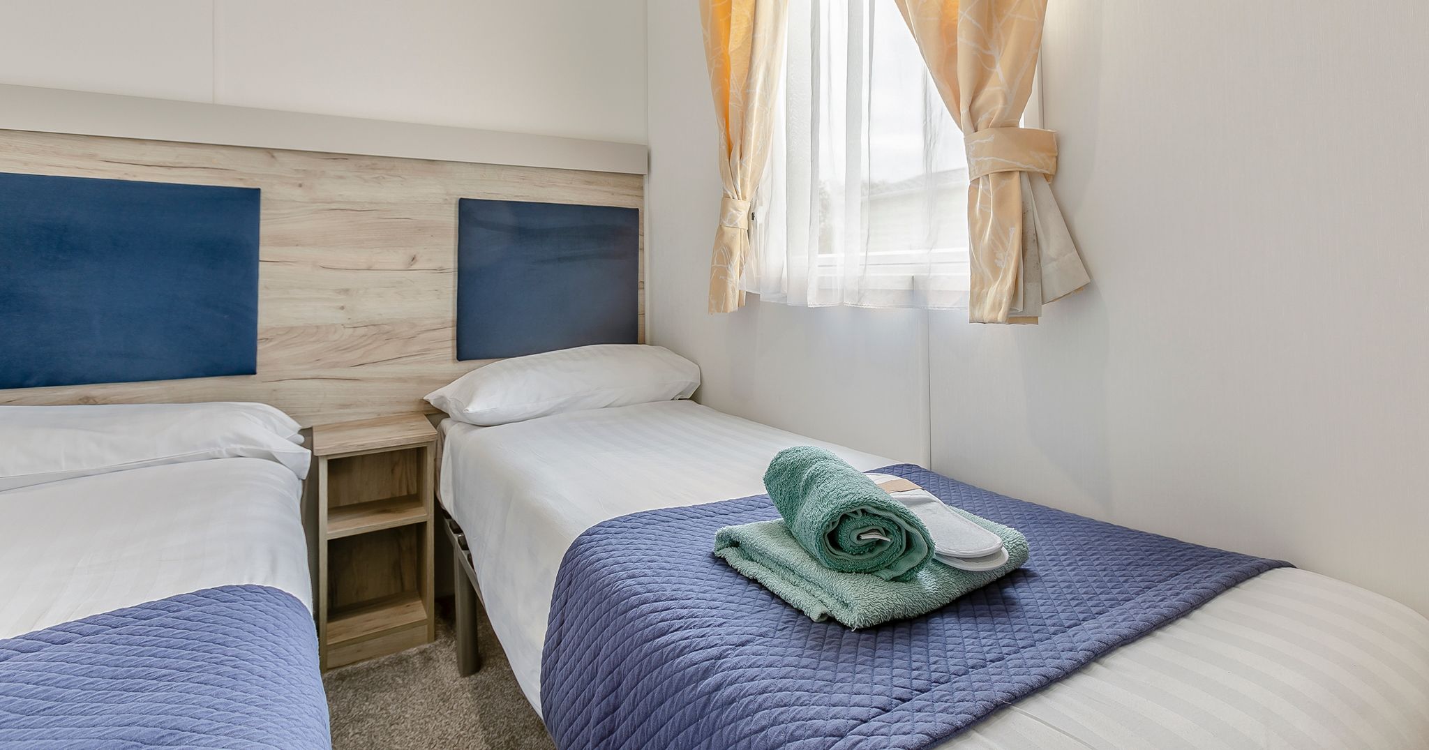 A tidy twin bedroom with two single beds, blue bed runners, folded towels on one bed, and a window with yellow curtains.