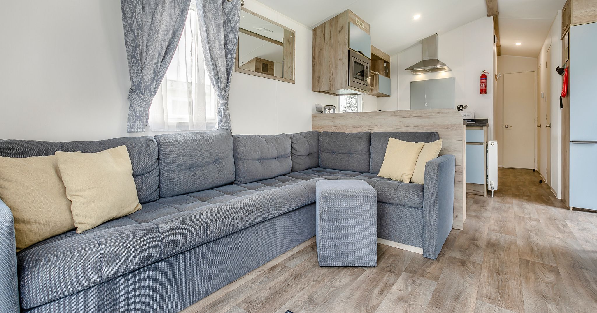 Modern living area with an L-shaped grey sofa, light wood floors, and open kitchen.