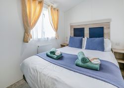 Bright and tidy bedroom with a double bed, blue bedding, rolled green towels, and slippers near a window with yellow curtains.