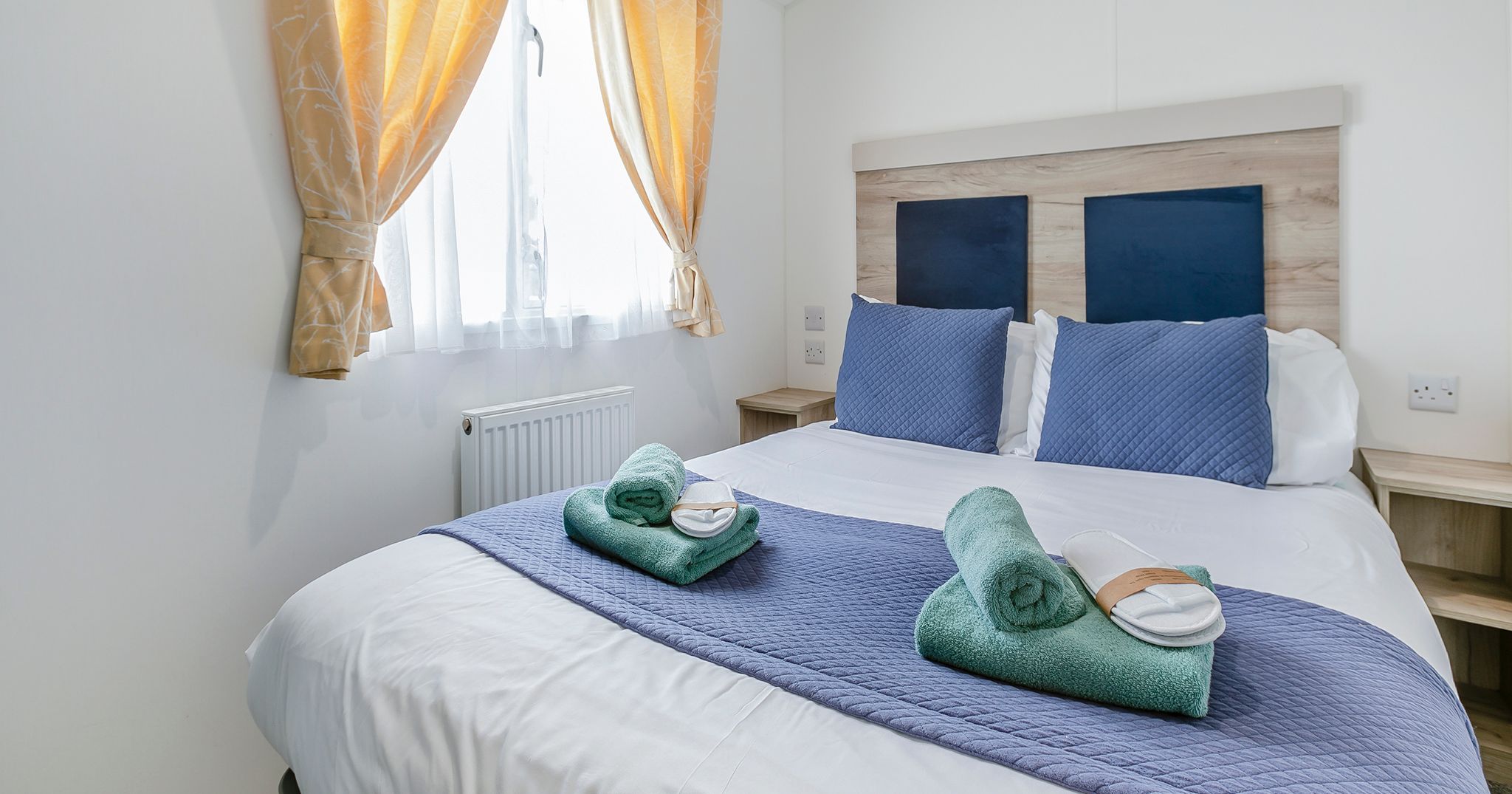 Bright and tidy bedroom with a double bed, blue bedding, rolled green towels, and slippers near a window with yellow curtains.