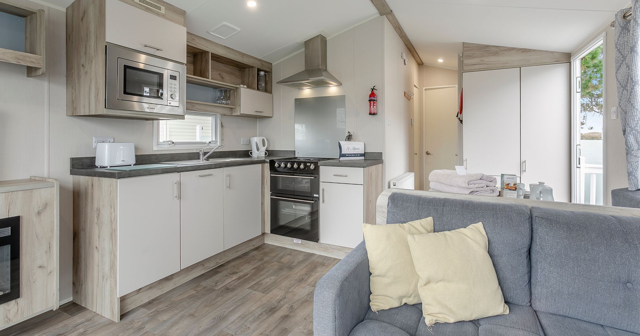 Modern kitchen and living area inside a mobile home with grey sofa and light wood flooring