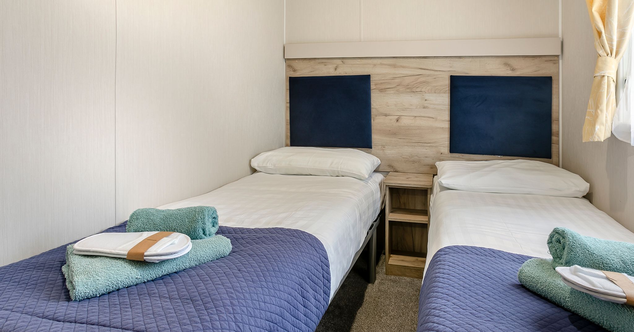 Twin beds in a small, modern room with blue covers and green towels neatly placed on each bed.