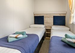 Twin beds in a small, modern room with blue covers and green towels neatly placed on each bed.