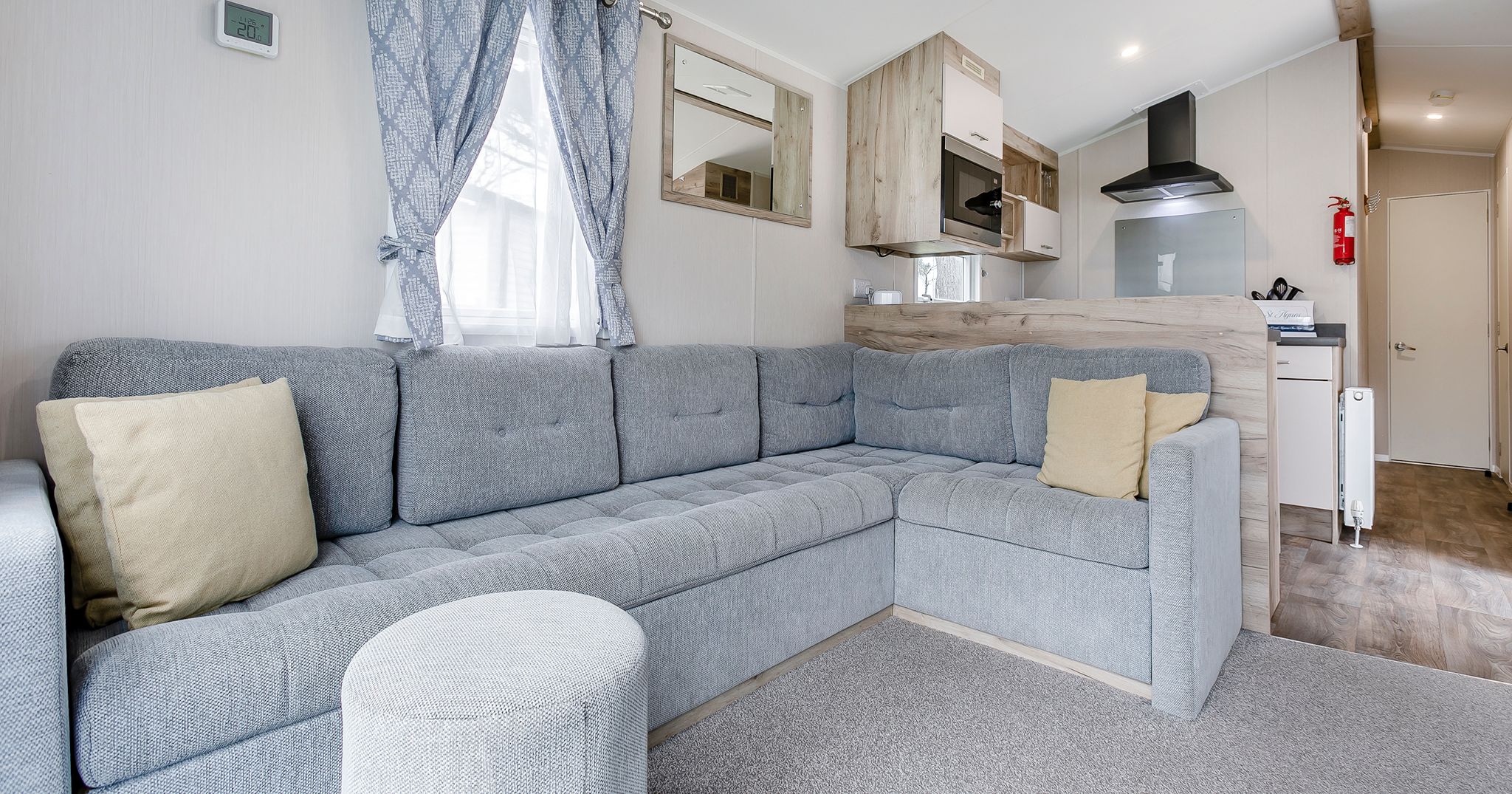 Modern living room with an L-shaped grey sofa, pillows, an ottoman, and a kitchen in the background.