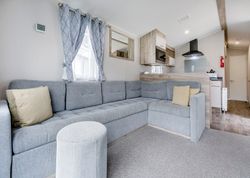 Modern living room with an L-shaped grey sofa, pillows, an ottoman, and a kitchen in the background.