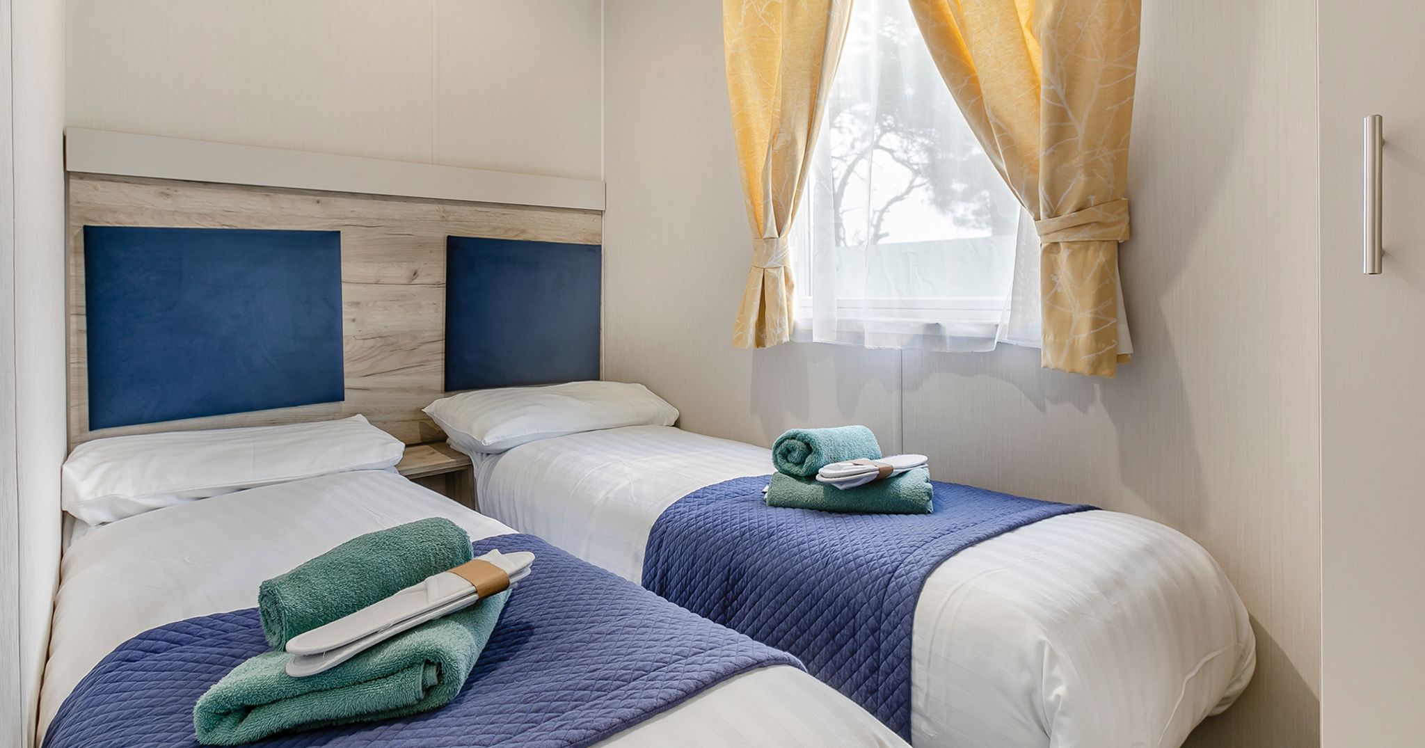 Two single beds with blue headboards, navy covers, and neatly arranged towels in a bright room with a window and yellow curtains.