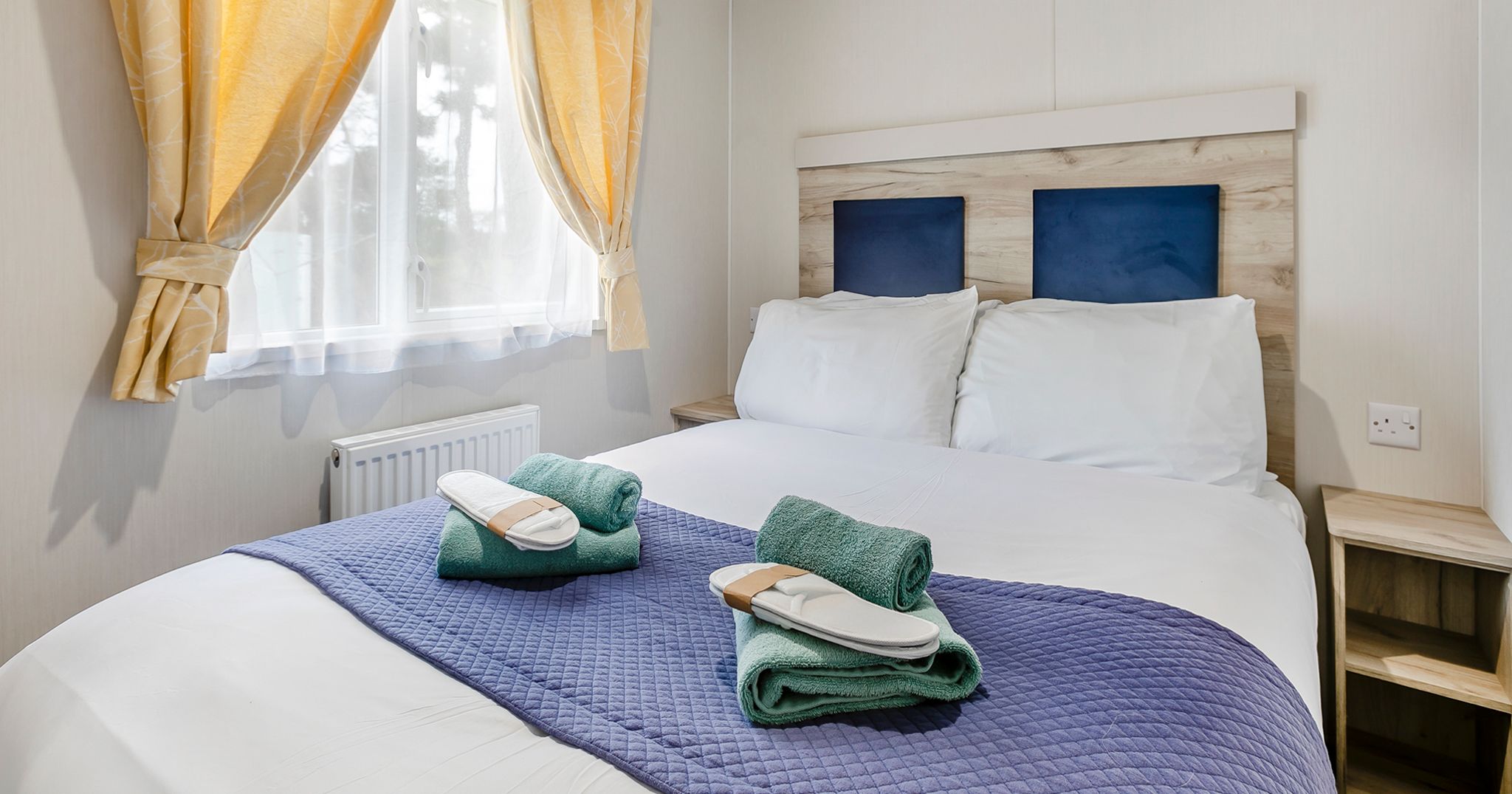 A neatly made double bed with white bedding, blue quilt, and two sets of green towels and white slippers. The room has yellow curtains and natural light coming through the window.