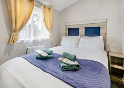 A neatly made double bed with white bedding, blue quilt, and two sets of green towels and white slippers. The room has yellow curtains and natural light coming through the window.